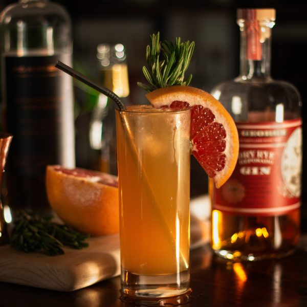 An orange garnish on a collins glass with bottles behind